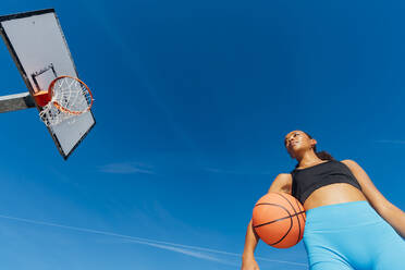 Nachdenklicher Basketballspieler mit Ball am Korb an einem sonnigen Tag - MEUF08189