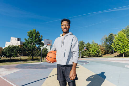 Selbstbewusster Spieler mit Basketball auf dem Platz an einem sonnigen Tag - MEUF08191