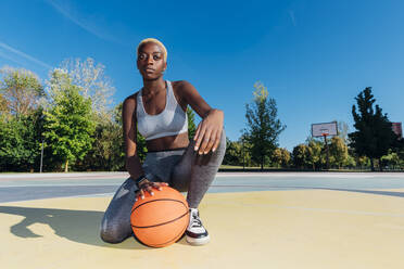 Selbstbewusste Sportlerin kniend mit Ball im Basketballfeld - MEUF08195