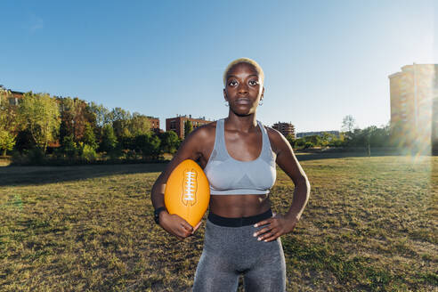 Junge Sportlerin mit Hand an der Hüfte, die einen American Football auf einem Feld hält - MEUF08250