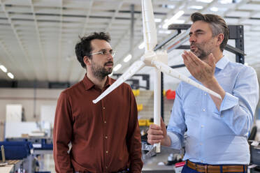 Mature businessman holding wind turbine discussing with colleague at warehouse - JOSEF13827