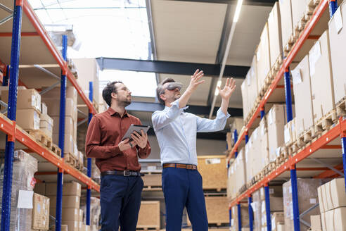 Businessman with colleague wearing virtual reality simulator gesturing at warehouse - JOSEF13836