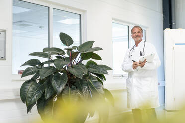 Smiling senior doctor with clipboard standing by potted plant - JOSEF13870