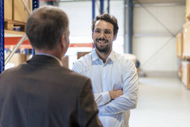 Happy businessman with arms crossed talking to colleague at warehouse - JOSEF13936