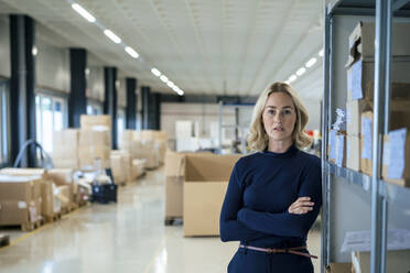 Confident businesswoman with arms crossed leaning on rack at warehouse - JOSEF13993