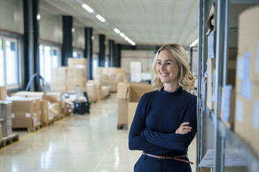 Happy blond businesswoman with arms crossed leaning on rack at warehouse - JOSEF13994