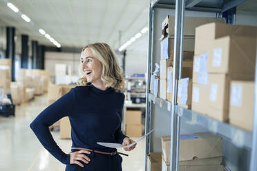 Cheerful businesswoman with document standing in front of rack at warehouse - JOSEF13996