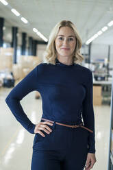 Smiling blond businesswoman with hand on hip standing at warehouse - JOSEF13997