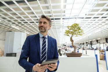 Smiling mature businessman with tablet PC standing in front of bonsai tree at industry - JOSEF14017