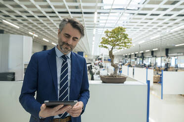Lächelnder reifer Geschäftsmann mit Tablet-PC vor einem Bonsaibaum in der Industrie stehend - JOSEF14018