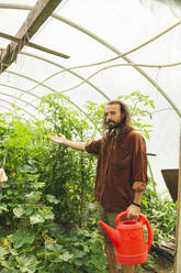 Junger Landwirt hält Gießkanne und gestikuliert bei Pflanzen im Gewächshaus - PCLF00099