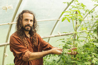 Smiling farmer with curly hair holding plant in greenhouse - PCLF00102