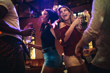 Glückliche junge Frauen tanzen und feiern mit männlichen Freunden in einem Nachtclub. Eine Gruppe von Freunden genießt an der Bar mit Getränken. - JLPSF04907