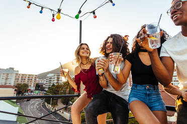 Junge Leute feiern auf der Terrasse mit Getränken. Männer und Frauen gemischter Rasse genießen Getränke auf dem Dach. - JLPSF04909
