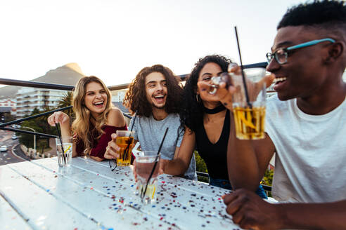 Eine multiethnische Gruppe von Freunden sitzt mit Getränken auf der Dachterrasse. Männer und Frauen feiern abends auf der Dachterrasse. - JLPSF04910