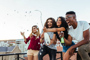 Freunde öffnen eine Flasche Champagner auf einer Dachterrasse. Junge Leute trinken Champagner auf einer Terrassenparty. - JLPSF04912