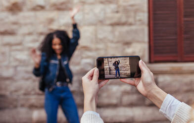 Woman clicking photo of her friend using mobile phone. Joyful woman posing for a photo while her friend uses mobile phone to capture the photo. - JLPSF04921