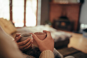 Nahaufnahme eines Paares, das einen Kaffee in den Händen hält, während es zu Hause auf dem Sofa sitzt, Fokus auf Hände und Kaffee. - JLPSF04926