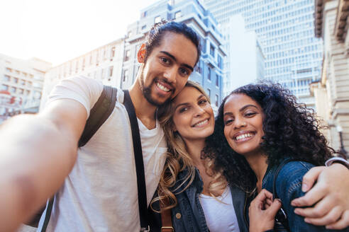 Cheerful man with two women friends clicking a selfie with city buildings in the background. Friends having fun while exploring the city. - JLPSF04927