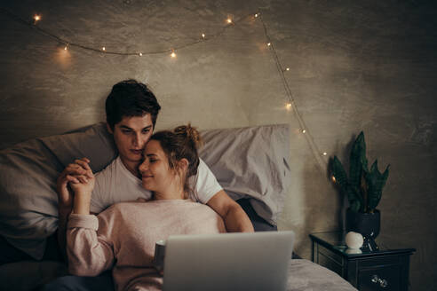 Romantisches junges Paar mit Laptop auf dem Bett. Verliebter Mann und verliebte Frau sehen sich ein Video auf dem Laptop an, während sie sich im Schlafzimmer entspannen. - JLPSF04931