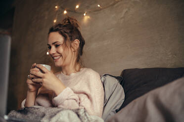 Fröhliche Frau trinkt Kaffee im Winter zu Hause. Kaukasische Frau entspannt sich in einem gemütlichen Schlafzimmer und trinkt Kaffee. - JLPSF04933
