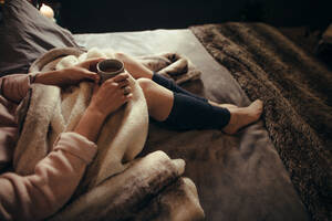 Ausschnitt einer Frau mit einer Tasse Kaffee auf dem Bett liegend, Fokus auf die Hände der Frau mit dem Kaffee. - JLPSF04935