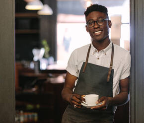 Nahaufnahme eines Barista, der am Eingang eines Coffeeshops steht und eine Kaffeetasse hält. Ein glücklicher Coffeeshop-Besitzer posiert mit einer Kaffeetasse in der Hand. - JLPSF04939
