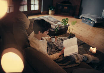 Junger Mann sitzt auf dem Sofa und liest ein Buch in einem gemütlichen Wohnzimmer. Mann entspannt sich im gemütlichen Wohnzimmer und liest ein Buch. - JLPSF04941