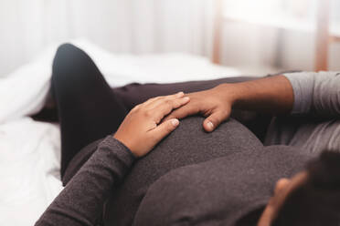 Nahaufnahme eines schwangeren Paares, das zu Hause auf dem Bett liegt; Fokus auf den Händen von Mann und Frau auf dem schwangeren Bauch. - JLPSF04988