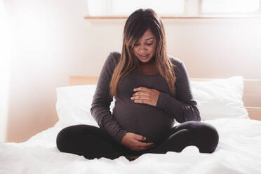 Beautiful pregnant woman sitting on bed at home. Young african pregnant woman relaxing in bedroom with her hands on her belly. - JLPSF04989