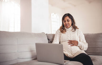 Portrait of beautiful young pregnant woman with laptop sitting on sofa. African woman expecting a baby sitting on couch and using laptop at home. - JLPSF04992