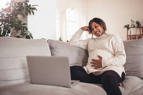 Porträt der glücklichen jungen schwangeren Frau, die auf dem Sofa sitzt und auf den Laptop schaut. Frau, die ein Baby erwartet, sitzt auf der Couch und benutzt den Laptop zu Hause. - JLPSF04993