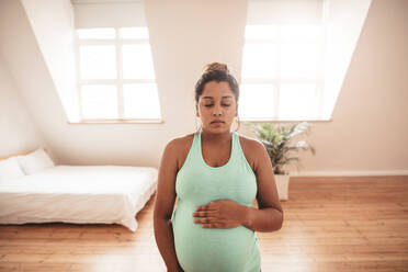 Porträt einer gesunden jungen schwangeren Frau, die im Wohnzimmer steht, ihren Bauch berührt und die Augen geschlossen hat. - JLPSF05000