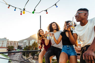 Vier junge Leute trinken und lächeln abends auf dem Dach. Männer und Frauen feiern mit Getränken auf der Terrasse. - JLPSF05006