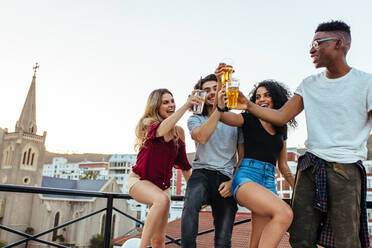 Freunde und Getränke. Junge, fröhliche Menschen, die auf einer Dachterrassenparty mit Bier anstoßen und lächeln. Männer und Frauen, die auf der Terrasse auf Getränke anstoßen. - JLPSF05011