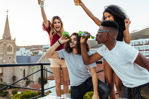 Gemischtrassige junge Leute feiern auf einer Terrasse, Männer trinken zusammen Bier und Mädchen jubeln. - JLPSF05013