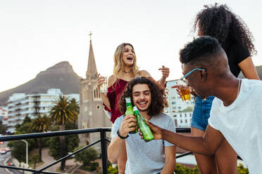 Glückliche junge Männer stoßen mit lachenden und genießenden Mädchen auf der Terrasse an. Eine Gruppe von Freunden feiert eine Dachparty mit Getränken. - JLPSF05014