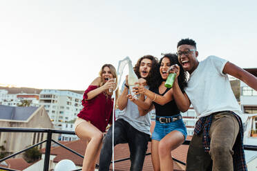 Cheerful friends celebrating rooftop party with champagne. Women opening the champagne bottle with her friends screaming and enjoying. Young men and women having a great time at the party. - JLPSF05016