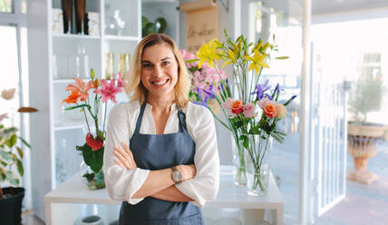 Beautiful and happy young woman florist standing in flower shop. Female florist in apron standing with her arms crossed and looking at camera. - JLPSF05080