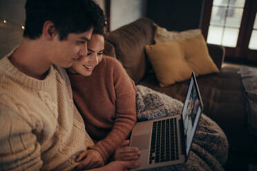 Mann und Frau sitzen auf dem Sofa und schauen auf einen Laptop im Wohnzimmer. Paar benutzt Laptop in hyggeligem Haus. - JLPSF05113