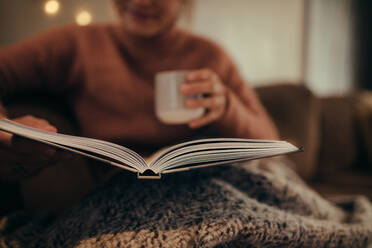 Nahaufnahme einer Frau, die mit einem aufgeschlagenen Buch und einem Kaffee auf dem Sofa sitzt und zu Hause ein Buch liest. - JLPSF05121