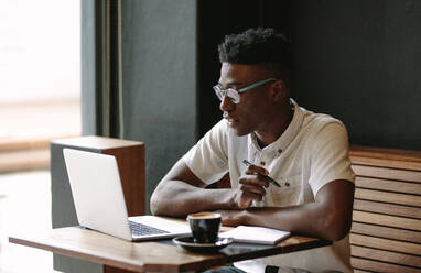 Ein Mann arbeitet an seinem Laptop in einem Café. Ein Freiberufler macht sich Notizen, während er an einem Kaffeetisch sitzt und auf seinen Laptop schaut. - JLPSF05123