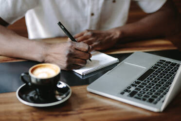 Freelancer writing notes sitting at a coffee table. Cropped shot of man working on his laptop computer at a coffee shop. - JLPSF05125