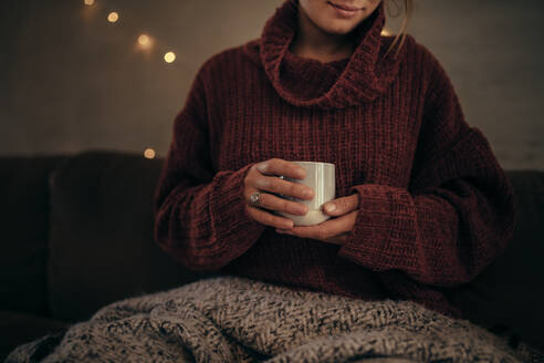 Nahaufnahme einer Frau, die im Winter zu Hause eine Tasse Kaffee in den Händen hält. Eine Frau entspannt sich im Wohnzimmer und trinkt Kaffee. - JLPSF05126