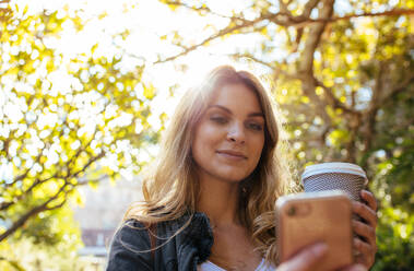 Frau schaut auf ihr Handy, während sie ein Kaffeeglas hält. Frau genießt Kaffee im Park, während sie ein Selfie mit dem Handy macht. - JLPSF05127