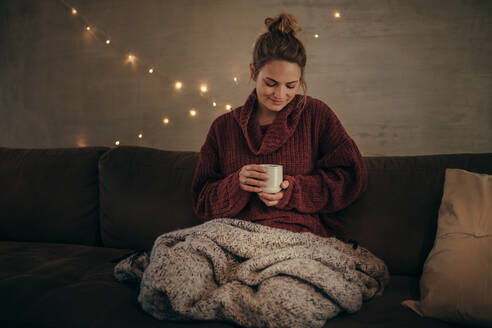 Entspannte Frau, die im Winter zu Hause Kaffee trinkt. Kaukasische Frau sitzt im gemütlichen Wohnzimmer mit einer Tasse Kaffee. - JLPSF05128
