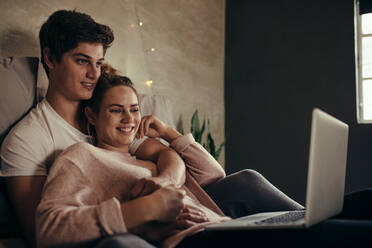 Gemütliches Pärchen mit Laptop auf dem Bett: Mann und Frau genießen es, ein Video auf dem Laptop anzuschauen, während sie sich im Schlafzimmer entspannen. - JLPSF05130