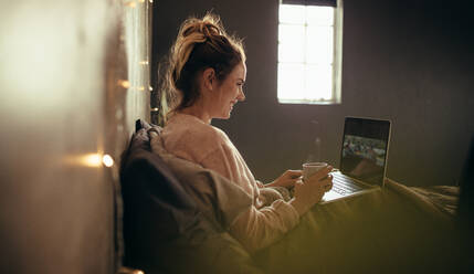Seitenansicht einer Frau auf dem Bett, die einen Laptop benutzt und Kaffee trinkt. Lächelnde Frau, die an einem Laptop arbeitet und zu Hause auf dem Bett Kaffee trinkt. - JLPSF05134