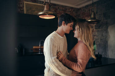 Seitenansicht eines jungen Mannes und einer verliebten Frau, die zusammen in einer Küche stehen. Romantisches junges Paar, das sich in der Küche umarmt. - JLPSF05143