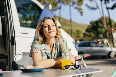 Fröhliche Frau, die ein Getränk mit Nachos genießt, während sie außerhalb des Wohnwagens auf dem Campingplatz in der Nähe von Surfbrett und Fahrrad sitzt - ADSF39350